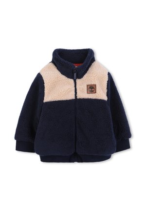 Veste en polaire bleu marine et beige à fermeture éclair avec un col haut, des poignets côtelés et un petit logo en patch marron sur la poitrine.