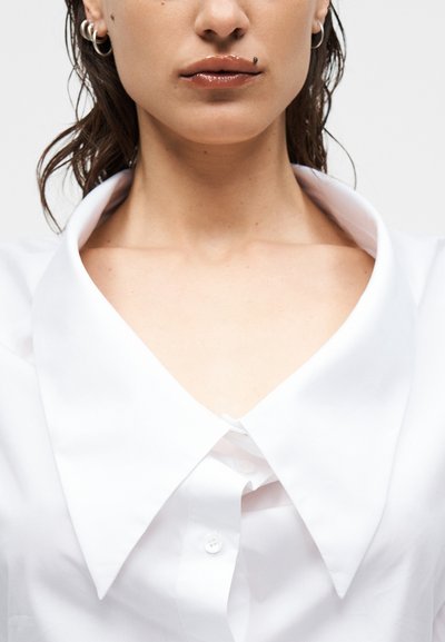 Γυναίκα που φοράει λευκό πουκάμισο με υπερμεγέθη μυτερό γιακά, λαμπερά χείλη, σημάδι ομορφιάς κοντά στο στόμα και πολλά ασημένια κρίκους σκουλαρίκια.