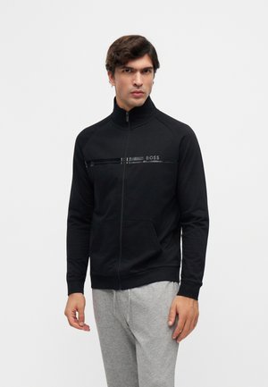 Hombre vestido con una chaqueta negra con cremallera y cuello alto con el logo "BOSS", combinado con pantalones deportivos gris claro, de pie contra un fondo liso.
