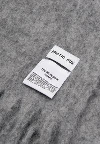 Écharpe grise en matériau doux et duveteux. Présente deux étiquettes blanches avec du texte noir indiquant la marque et les instructions d'entretien.