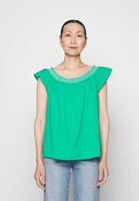 Top verde com um design amplificado, apresentando um decote decorativo bordado e um tecido suave e leve. Combinado com jeans azuis.