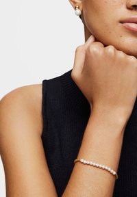 Pendientes de perlas en tono dorado y una pulsera delicada con pequeñas cuentas doradas y perlas blancas alternadas, llevados en un brazo desnudo.