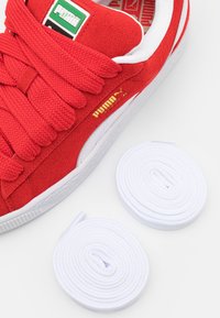 Red suede sneaker with white accents and bold laces. Includes two white round shoe laces. Gold logo on the side.