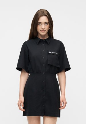 Pruunide pikkade juustega naine, kes kannab musta nööbitavat kleiti, millel on laiad lühikesed varrukad ja rinnatasku valge tekstiga.