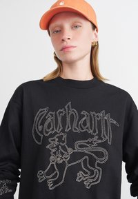 Crna dukserica sa složenim bijelim vezom lava i natpisom "Carhartt". Uparen s narančastom kapom.