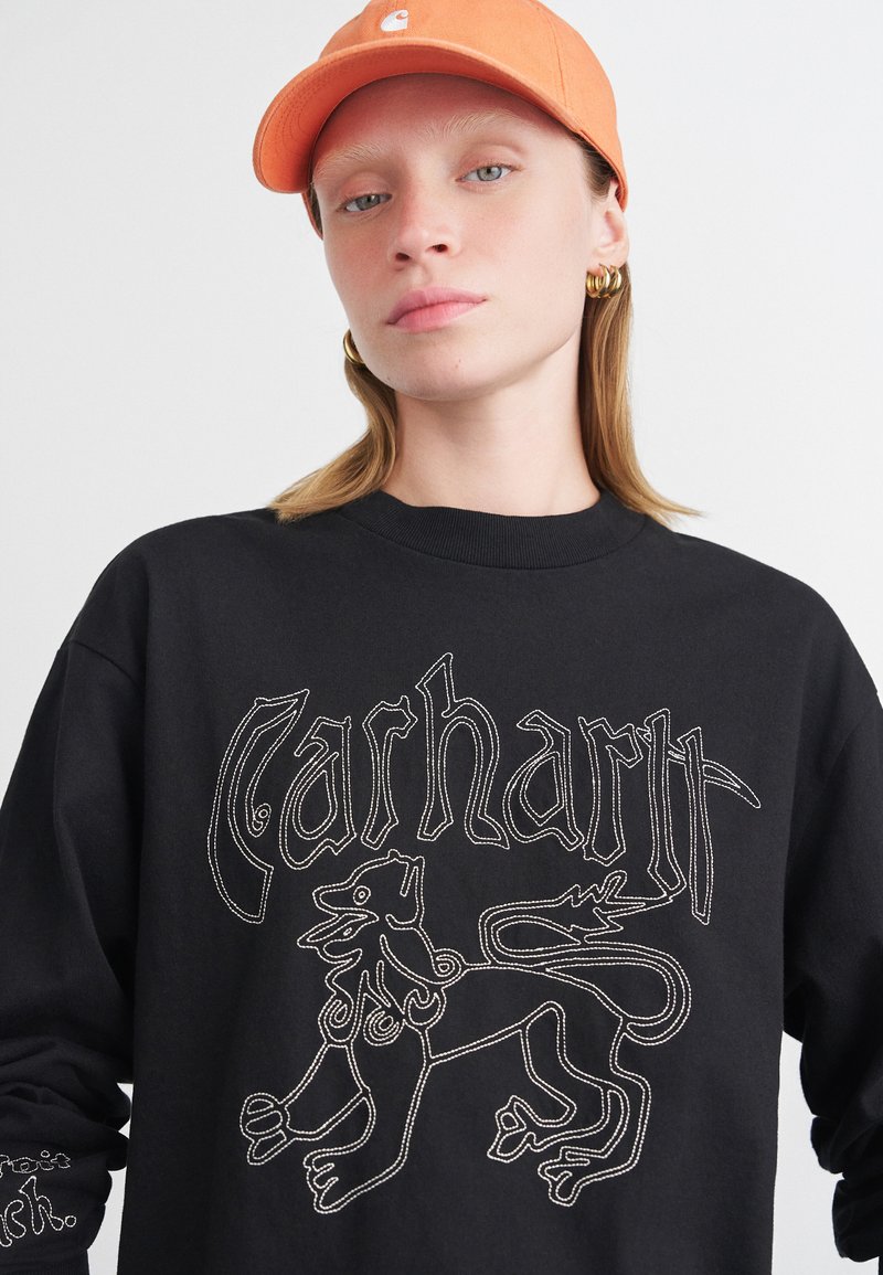 Crna dukserica sa složenim bijelim vezom lava i natpisom "Carhartt". Uparen s narančastom kapom.
