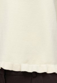 Pull en maille blanc cassé avec un ourlet à volants subtil, associé à un pantalon marron foncé.