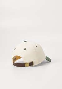 Baseball cap med en cremefarvet bomuldsdel og mørkegrøn skygge. Brun justerbar rem bagpå med en messing spænde og logo detalje.