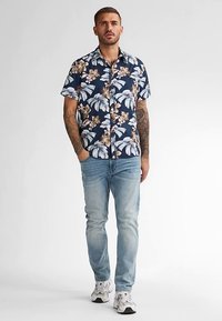 Korte mouwen, marineblauw overhemd met bloemenpatroon in lichtblauw en beige, gecombineerd met lichtblauwe jeans en witte sneakers.