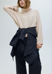 Bézs gyapjú garbó sötét csíkos anyag fölött viselve, deréknál összehúzva, laza fekete nadrággal és kéz kiegészítővel.