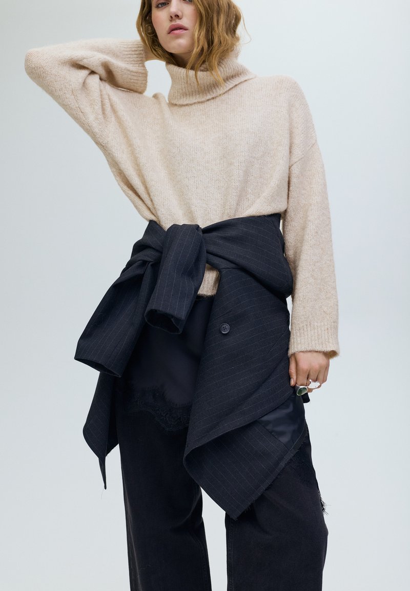 Bézs gyapjú garbó sötét csíkos anyag fölött viselve, deréknál összehúzva, laza fekete nadrággal és kéz kiegészítővel.