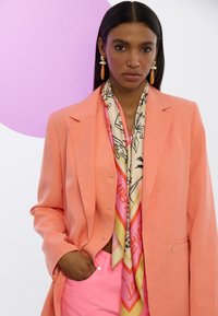 Korallblazer mit lässiger Passform, kombiniert mit einem bunten gemusterten Seidenschal und rosa Hosen. Die Ohrringe haben einen runden orangen Akzent.