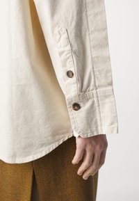 Långärmad beige skjorta i bomull med knäppta ärmslut. Har en spetsig ärmpaket och en slät yta. Handen vilar nära fållen.