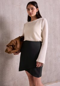 Krémový pletený svetr s dlouhými rukávy, doplněný černou zavinovací sukní. Modelka drží hnědou velkou tašku.