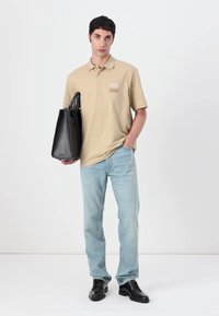 Μπεζ πουκάμισο πόλο με μικρή τσέπη, σε συνδυασμό με ανοιχτό μπλε τζιν και μαύρες μπότες. Το μοντέλο κρατά μια μαύρη τσάντα tote.