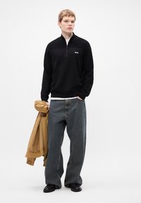 Μαύρο φούτερ με φερμουάρ και υφή στην επάνω πλευρά, λευκό λογότυπο, χαλαρές γκρίζες τζιν παντελόνες και μια καμηλό μπόμπερ τζάacket κρεμασμένη στον βραχίονα. Μαύρα παπούτσια.