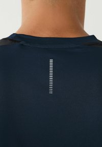 Marineblå atletisk shirt med en glat tekstur, der har en vertikal reflekterende detalje på ryggen og kontrasterende sorte accenter på skuldrene.