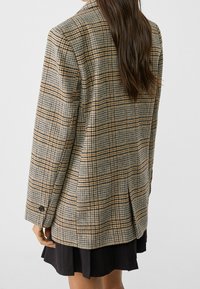 Blazer à carreaux en gris, marron et beige, avec une coupe ajustée, des manches longues et des boutons décoratifs aux poignets.