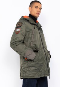 Zielona pikowana kurtka z pomarańczową podszewką, odpinanym kapturem, wieloma kieszeniami, szarymi akcentami i haftowanymi naszywkami na rękawach.