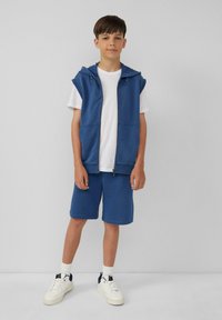 Niebieska bezrękawnik z kapturem, zapinany na zamek, noszony na białej koszulce, w połączeniu z niebieskimi szortami i białymi trampkami. Prosty design z casualowym krojem.