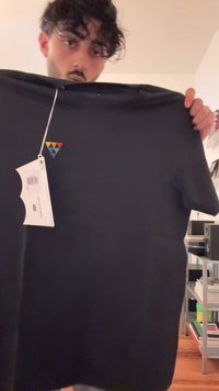 Jauns vīrietis ar tumšiem cirtainiem matiem tur melnu t-kreklu ar mazu krāsainu trīsstūra logo un pievienotu cenu zīmi.
