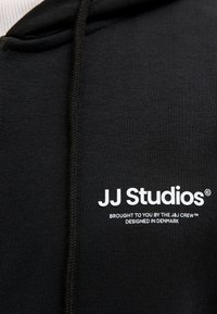 Hoodie noir avec un logo graphique blanc "JJ Studios" accompagné de détails textuels. Tissu doux et coupe régulière soulignés par une capuche avec cordon.