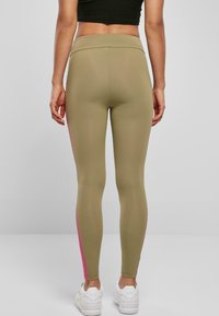 Olivenfargede leggings med en tettsittende design, med en rosa stripe langs den ytre sømmen. Laget av et elastisk materiale. Hvite sneakers synlige.