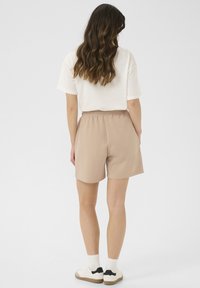 Pantaloni corti in cotone beige con vita elastica, tasche laterali e una vestibilità rilassata, abbinati a una maglietta bianca a maniche corte e scarpe da ginnastica.