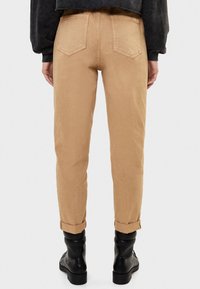 Pantalones en tono beige, ajustados y con puños enrollados, fabricados con un tejido resistente. Cuentan con dos bolsillos traseros y una cintura de altura media. Combinados con botas negras de tobillo.