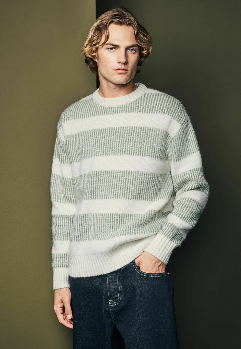 Knit sweater with green and white stripes, round neck, and ribbed texture. Paired with dark denim pants.