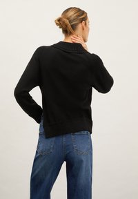 Svart stickad tröja med ribbat krage och manchetter, tillsammans med blå denimjeans med en traditionell femfickdesign och synlig sömnad.