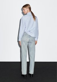 Camicia a righe blu chiaro con maniche lunghe infilata in jeans dritti sbiaditi, abbinata a scarpe nere. Vista posteriore visibile.