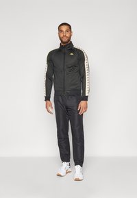 Black athletic jacket with gold accents, full zip, high collar, and patterned sleeves. Worn with dark pants and white sneakers.