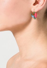 Personne portant une boucle d'oreille en or ornée de pierres multicolores en forme de feuilles sur l'oreille gauche, sur un fond uni.