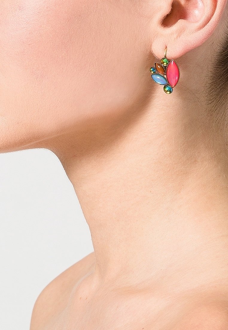 Personne portant une boucle d'oreille en or ornée de pierres multicolores en forme de feuilles sur l'oreille gauche, sur un fond uni.
