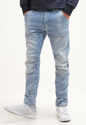 Mann trägt hellblaue, ausgewaschene Jeans mit Knie-Nähdetails und weißen Turnschuhen, Hände in den Taschen vor einem einfarbigen Hintergrund.