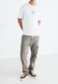 Bílá bavlněná tričko s grafickým potiskem "Santorini" a béžové cargo kalhoty s několika kapsami, doplněné hnědými a krémovými teniskami.