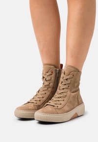 Beige high-top sneakers with suede material, textured surface, rubber sole, front lacing, and zip closure detail on the side.