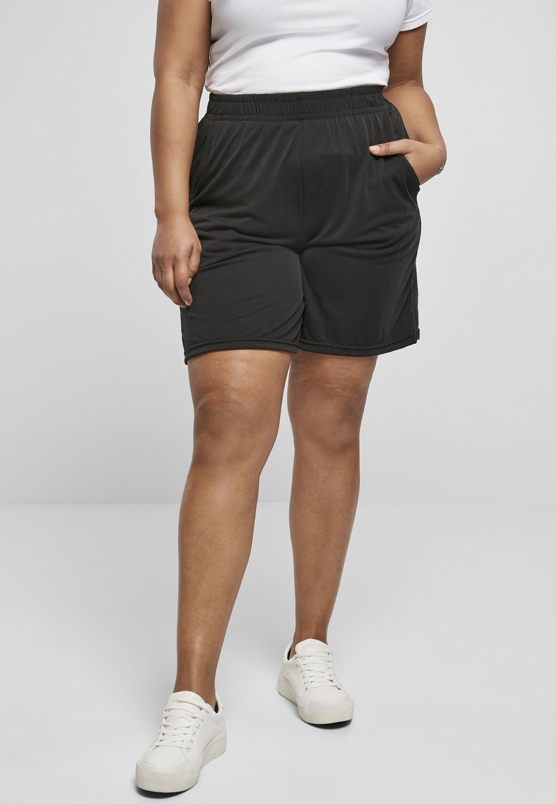 Sorte treningshorts med elastisk midjebånd, sidelommer og løs passform. Brukt med hvite joggesko mot en nøytral bakgrunn.