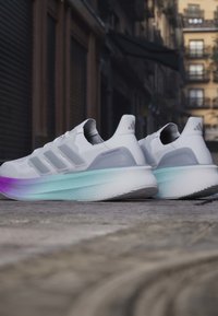 Λευκά παπούτσια τρεξίματος Adidas Ultraboost με ελαφρώς μοβ και τιρκουάζ υποσόλους σε διαβάθμιση, τοποθετημένα σε λιθόστρωτο δρόμο σε αστικό περιβάλλον.