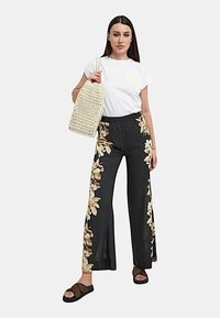 T-shirt bianco con una toppa floreale, pantaloni larghi neri a fantasia con dettagli floreali, borsa intrecciata beige, e sandali neri.