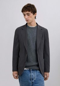 Blazer gris con solapas con muesca, llevado sobre un suéter gris texturizado. Jeans azules completan el conjunto. Diseño simple y moderno.