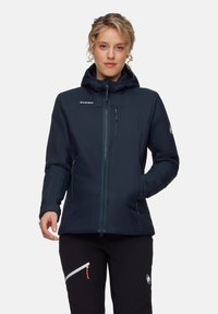 Kvinna i en mörkblå Mammut jacka med huva, som har zippered bröstfickor och sidfickor, tillsammans med svarta byxor som har en zippered ficka.