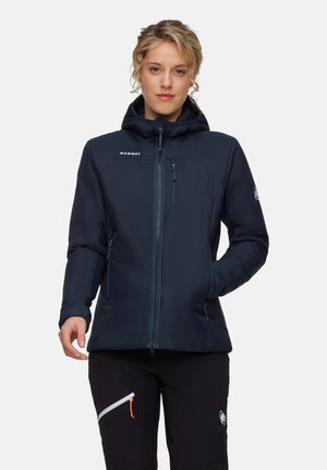Donna che indossa una giacca con cappuccio Mammut di colore blu navy, dotata di tasche con zip sul petto e sui lati, abbinata a pantaloni neri con una tasca con zip.