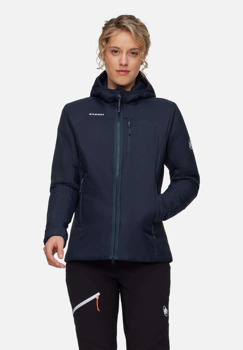 Kvinna i en mörkblå Mammut jacka med huva, som har zippered bröstfickor och sidfickor, tillsammans med svarta byxor som har en zippered ficka.