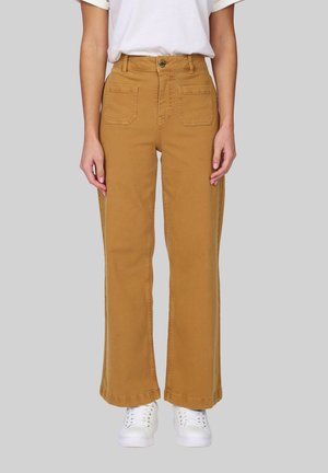 Personne portant un pantalon moutarde taille haute à jambes larges avec poches plaquées à l'avant et des baskets blanches, debout devant un fond gris uni.