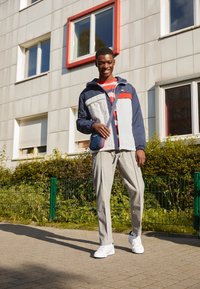 Veste pour homme bicolore en bleu marine, blanc et rouge ; chemise rayée rouge et blanche ; pantalon gris clair ; baskets blanches ; petit sac bandoulière bleu.