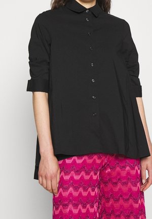 Frau trägt ein lockeres, schwarzes Hemd mit aufgekrempelten Ärmeln und glänzende, pinke Hose mit Zickzackmuster vor einem schlichten Hintergrund.