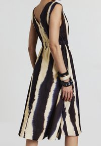 Ärmlös midiklänning med beige och svarta vertikala batikrandningar, elastisk midja, djup V-nacke, bärs med svart och vitt armband på vänster handled.