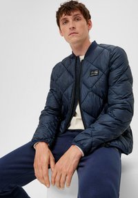 Navyblau gesteppte Bomberjacke mit Reißverschluss auf der Vorderseite, geripptem Kragen und Logo-Patch. Über einem hellen Hemd getragen, kombiniert mit dunklen Hosen.
